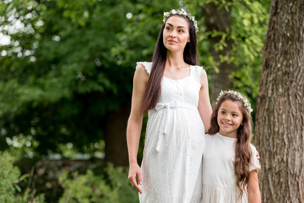 onnellinen raskaana äiti ja söpö tytär valkoisissa mekoissa ja seppeleissä seisoo yhdessä ja katselee pois puistossa
 - Valokuva, kuva