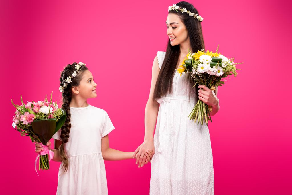 ομορφη ευτυχισμένη μητέρα και κόρη σε άσπρα φορέματα και floral στεφάνια κρατώντας ανθοδέσμες και χαμογελαστά κάθε άλλο απομονώνονται σε ροζ - Φωτογραφία, εικόνα