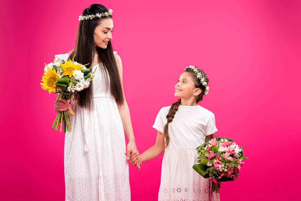 όμορφη μητέρα και κόρη στην άσπρα φορέματα κρατώντας ανθοδέσμες και χαμογελαστά κάθε άλλο απομονώνονται σε ροζ - Φωτογραφία, εικόνα