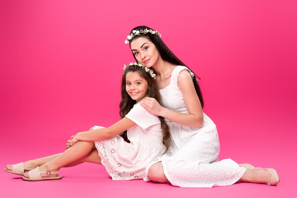 Вид в полный рост красивых счастливых матери и дочери в белых платьях и цветочных венках, сидящих вместе и улыбающихся на камеру в розовом
 - Фото, изображение