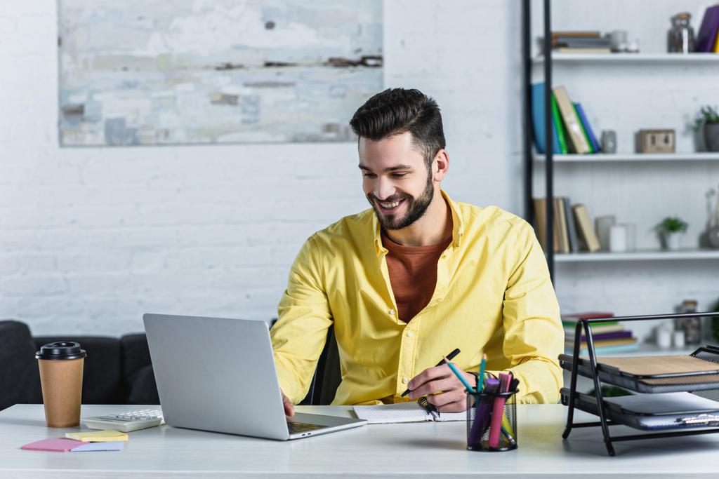 Empresário de camisa amarela segurando caneta e olhando para a tela do laptop
 - Foto, Imagem