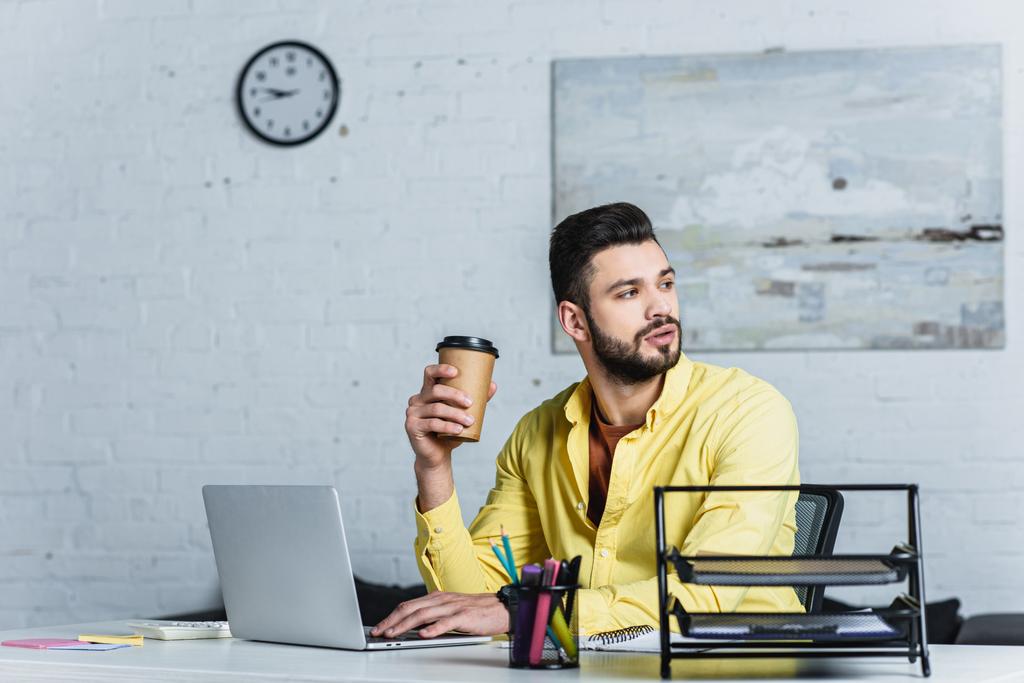 Bearded businessman holding disposable cup and looking away at workplace - Photo, Image