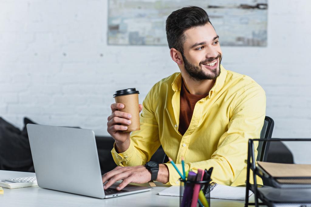 Hombre de negocios sonriente sosteniendo una taza de papel, usando un portátil y mirando hacia otro lado
 - Foto, Imagen