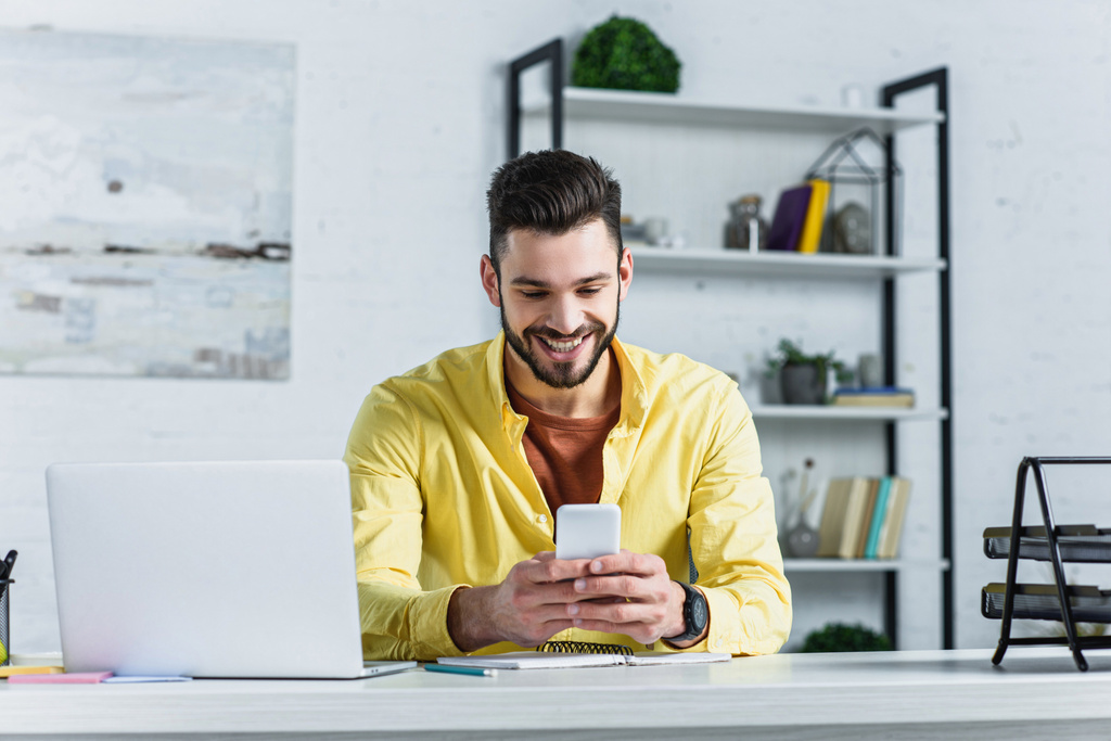 Усміхнений бородатий бізнесмен в жовтій сорочці дивиться на смартфон на робочому місці
 - Фото, зображення