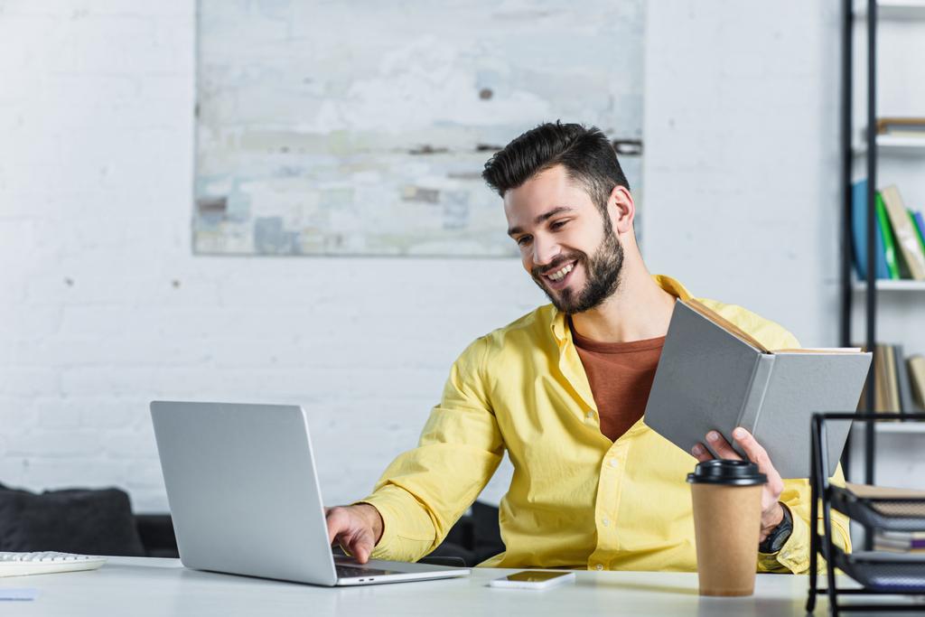 Hymyilevä parrakas liikemies katselee kannettavaa tietokonetta ja pitää kirjaa työpaikalla
 - Valokuva, kuva