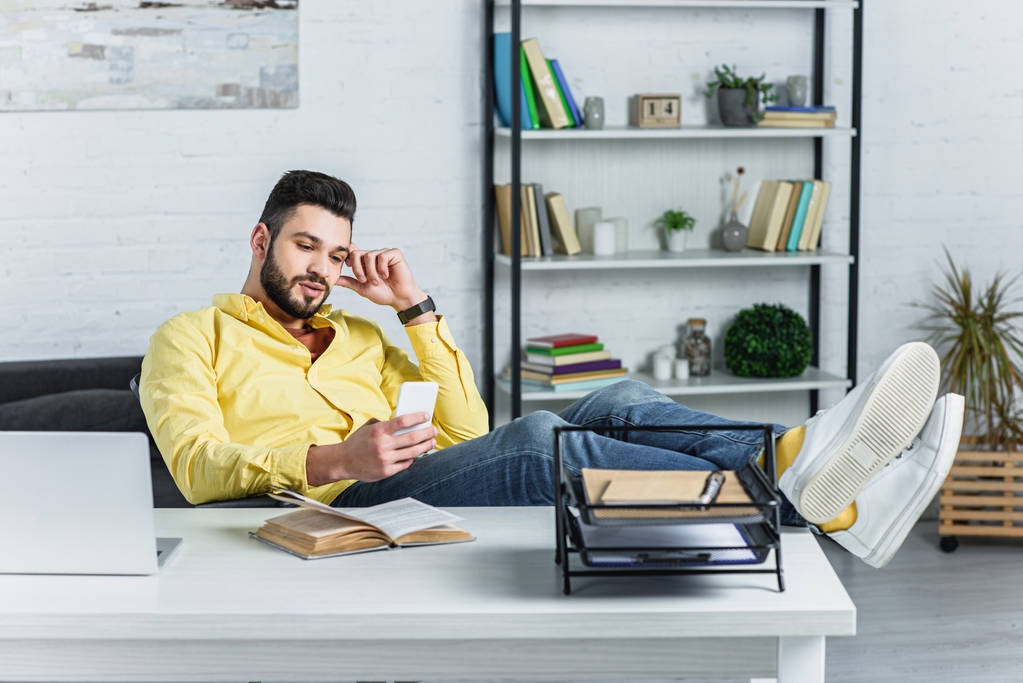 Empresário barbudo pensativo em camisa amarela olhando para o smartphone no local de trabalho
 - Foto, Imagem