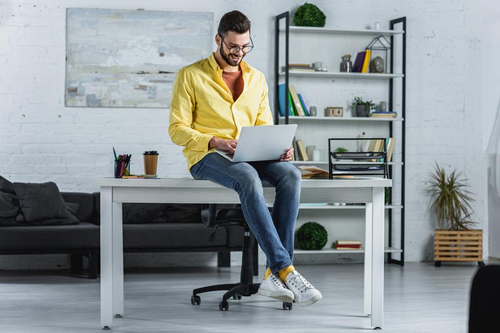 Χαμογελώντας γενειοφόρος επιχειρηματία σε ποτήρια πληκτρολογώντας στο laptop και να κάθεται στο γραφείο - Φωτογραφία, εικόνα