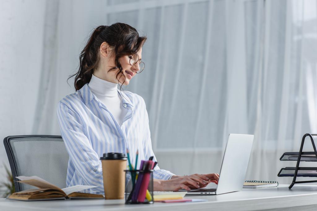 Empresária alegre em óculos digitando no laptop no escritório
 - Foto, Imagem
