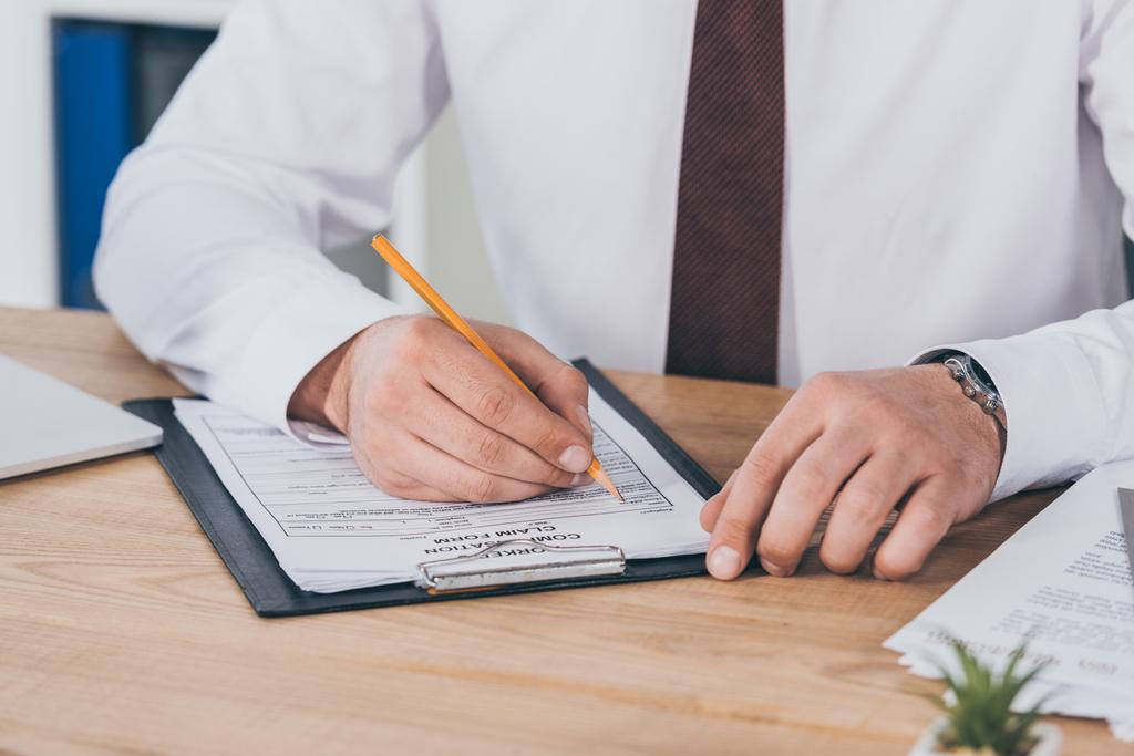 cropped view of businessman writing on compensation claim form at workplace - Photo, Image