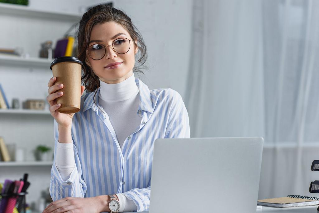 Femme d'affaires attrayante dans des lunettes tenant tasse en papier et regardant loin sur le lieu de travail
 - Photo, image