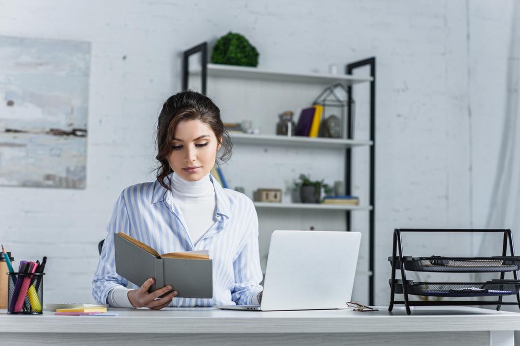 mulher atraente estudando com livro perto de laptop no escritório moderno
  - Foto, Imagem