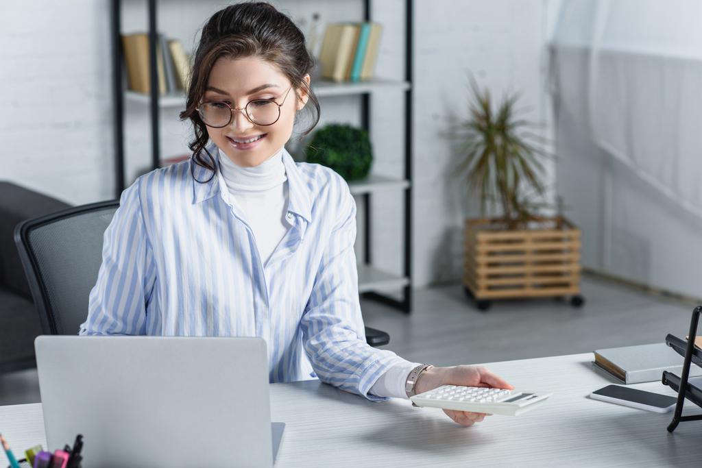 attraktive Frau mit Brille, Taschenrechner in der Nähe von Laptop im modernen Büro  - Foto, Bild