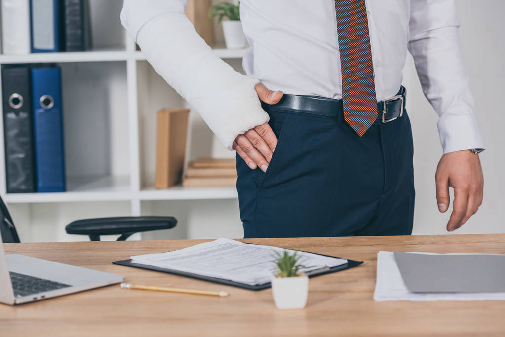 cropped view of worker with broken arm holding hand in pocket and standing at table in office, compensation concept - Photo, Image
