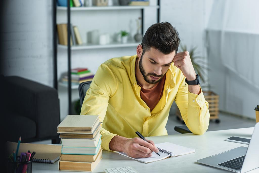 focused man writing in notebook near laptop and books in modern office  - Photo, Image