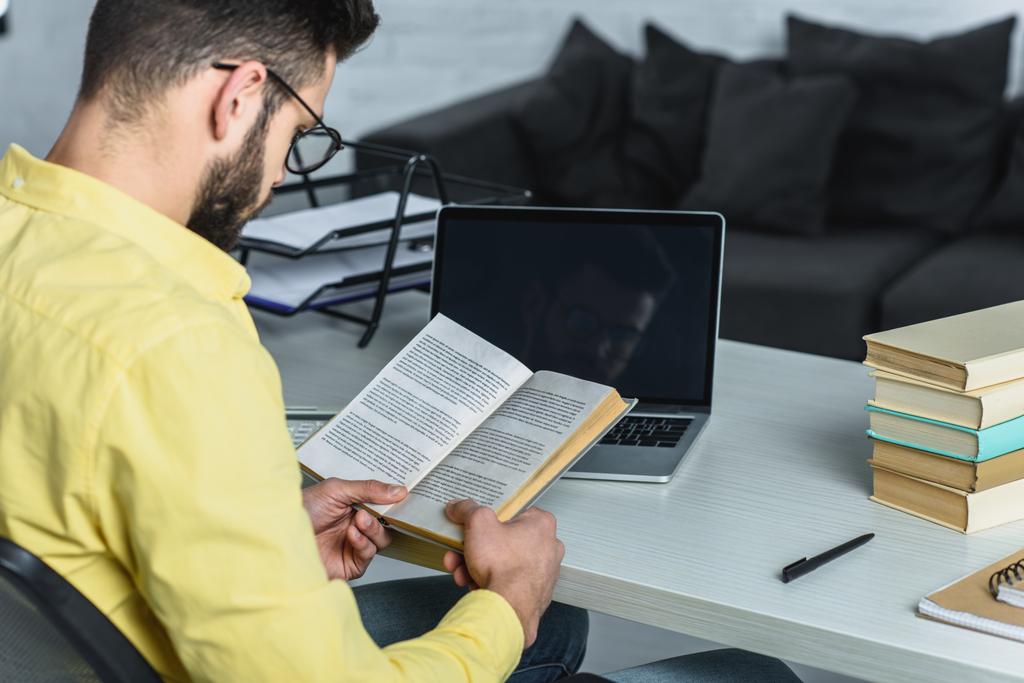 bebaarde man studeren met boek in de buurt van laptop met leeg scherm in moderne kantoren - Foto, afbeelding