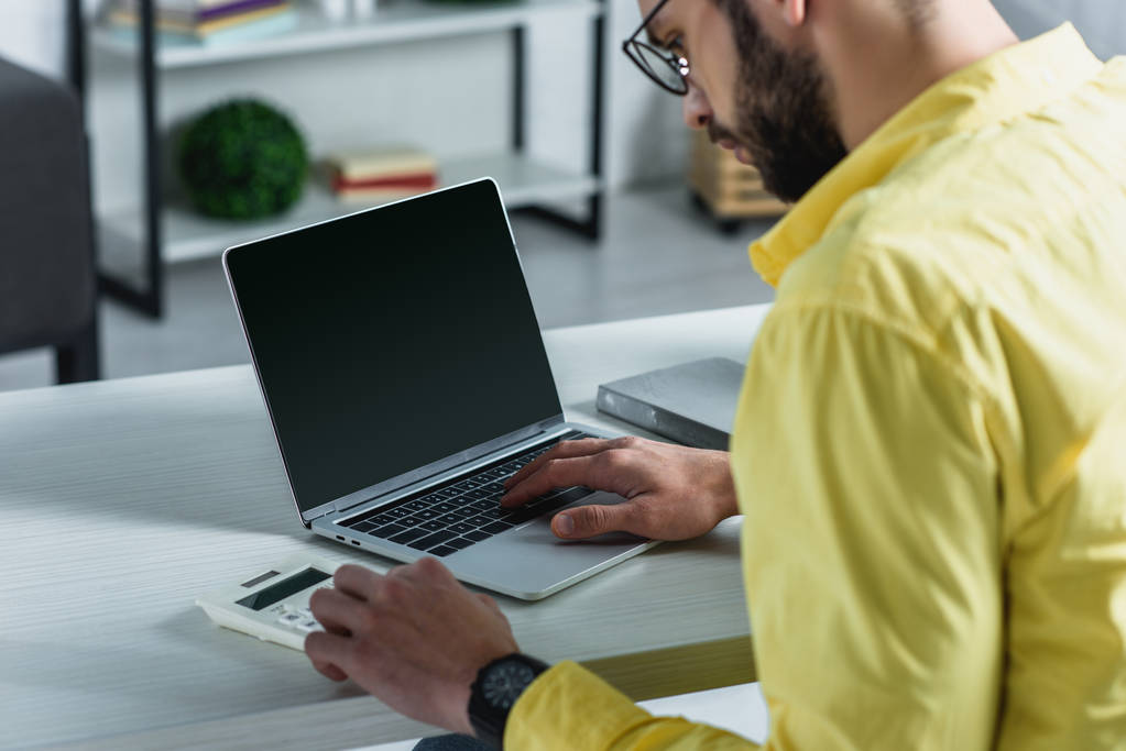 bärtiger Mann blickt in modernem Büro auf Taschenrechner neben Laptop mit leerem Bildschirm - Foto, Bild