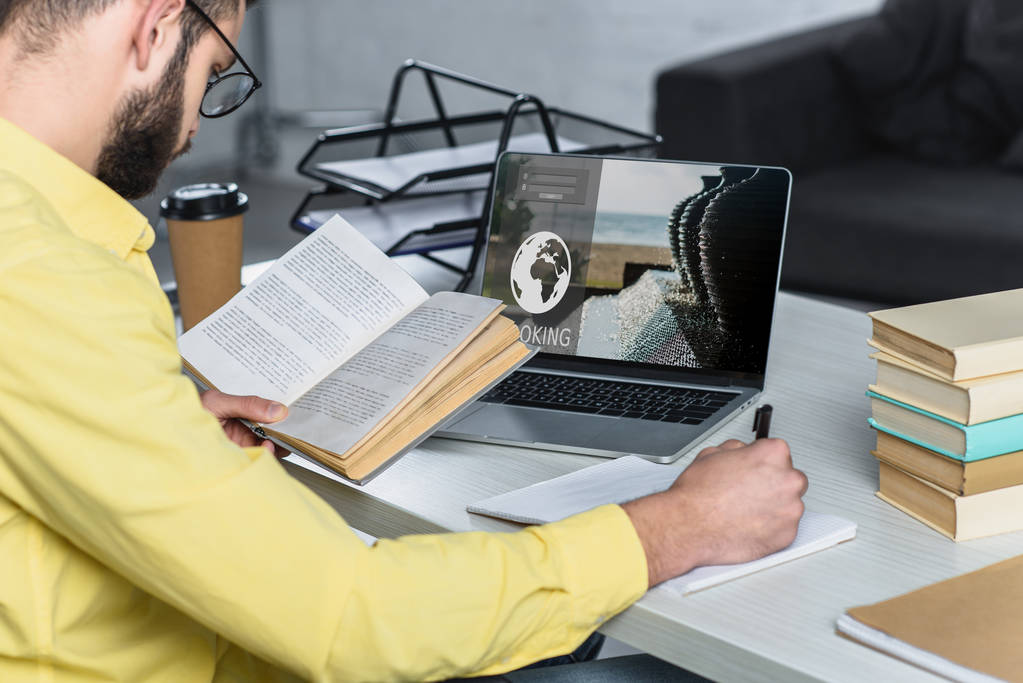 homme barbu étudiant avec livre près d'un ordinateur portable avec site de réservation à l'écran dans le bureau moderne
 - Photo, image