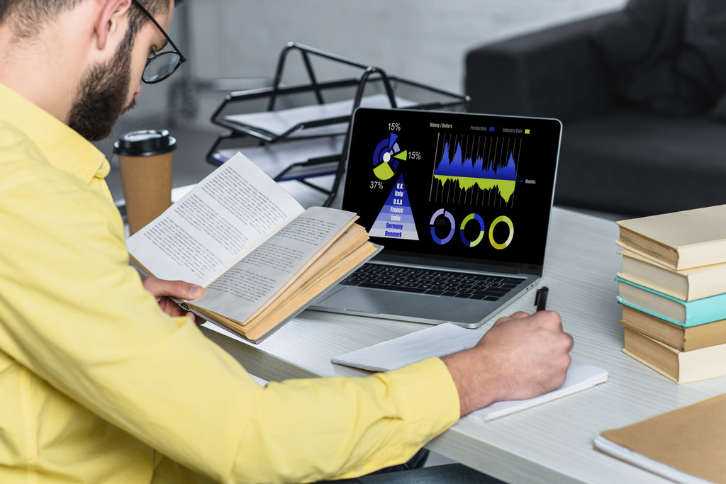 бородатий чоловік навчається з книгою біля ноутбука з графіками і графами на екрані в сучасному офісі
 - Фото, зображення
