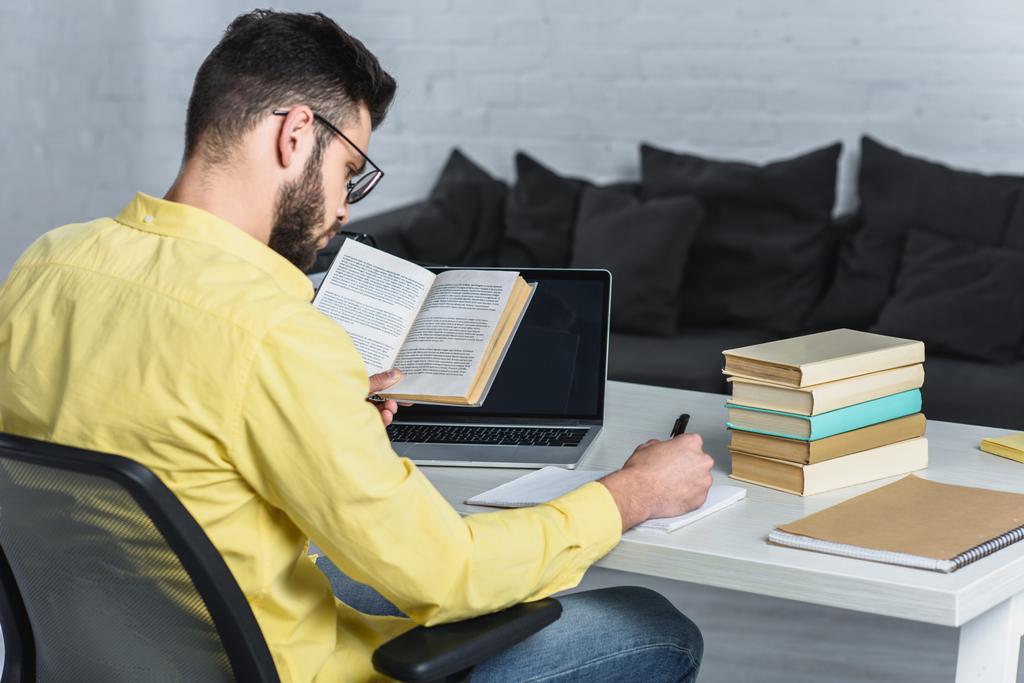 γενειοφόρος άνδρας, μελετώντας με βιβλίο κοντά σε φορητό υπολογιστή με κενή οθόνη στο σύγχρονο γραφείο - Φωτογραφία, εικόνα