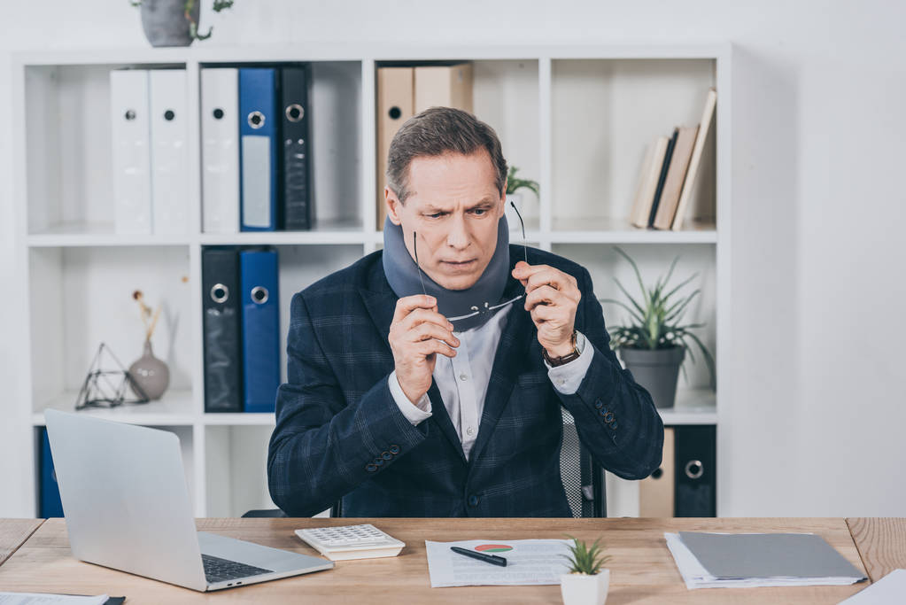 бизнесмен в шейном корсете сидит за столом с документами, накладывая на очки в офисе, концепция компенсации
 - Фото, изображение