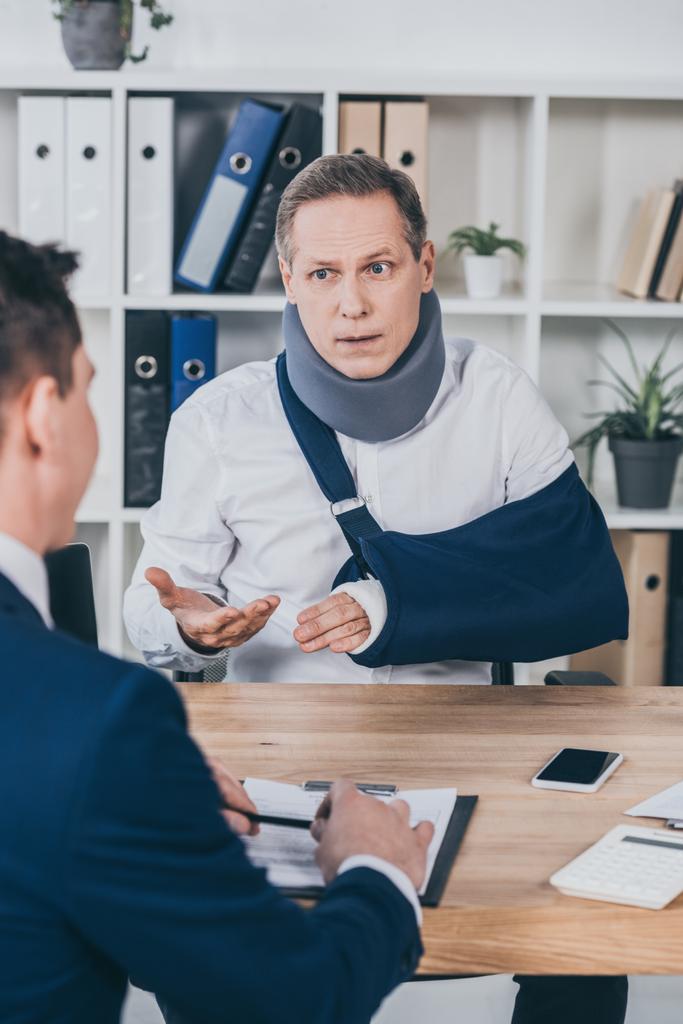 środkowy wieku pracownika w ortopedyczny ze złamaną rękę, siedzi przy stole i rozmawia z przeciwnej biznesmen w biurze, pojęcie odszkodowania - Zdjęcie, obraz