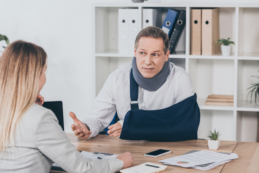 middle aged worker in neck brace with broken arm sitting at table and talking to blond woman in office, compensation concept - Photo, Image
