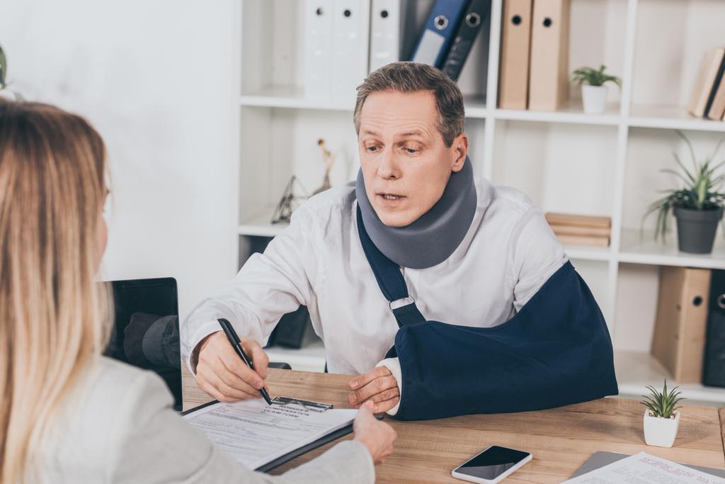 worker in neck brace and arm bandage at table pointing with pen in document while woman sitting opposite in office, compensation concept - Photo, Image