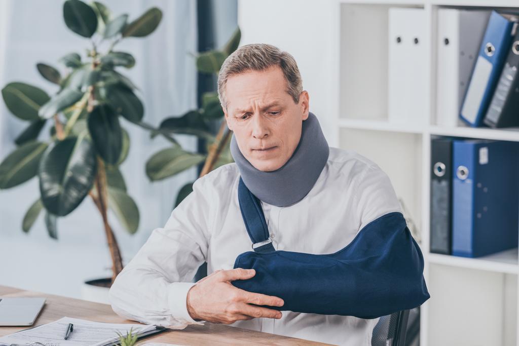 trabalhador de meia-idade segurando braço quebrado wile sentado à mesa com documento no escritório, conceito de compensação
 - Foto, Imagem