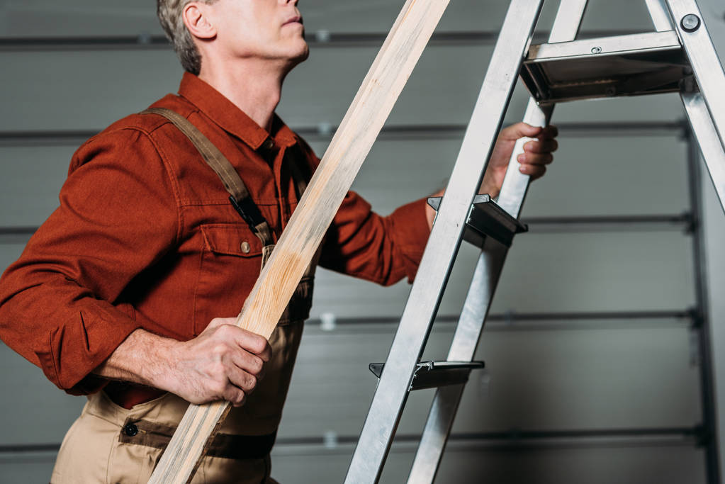 cropped view of repairman in orange uniform climbing with wooden board in hand on ladder in garage - Photo, Image