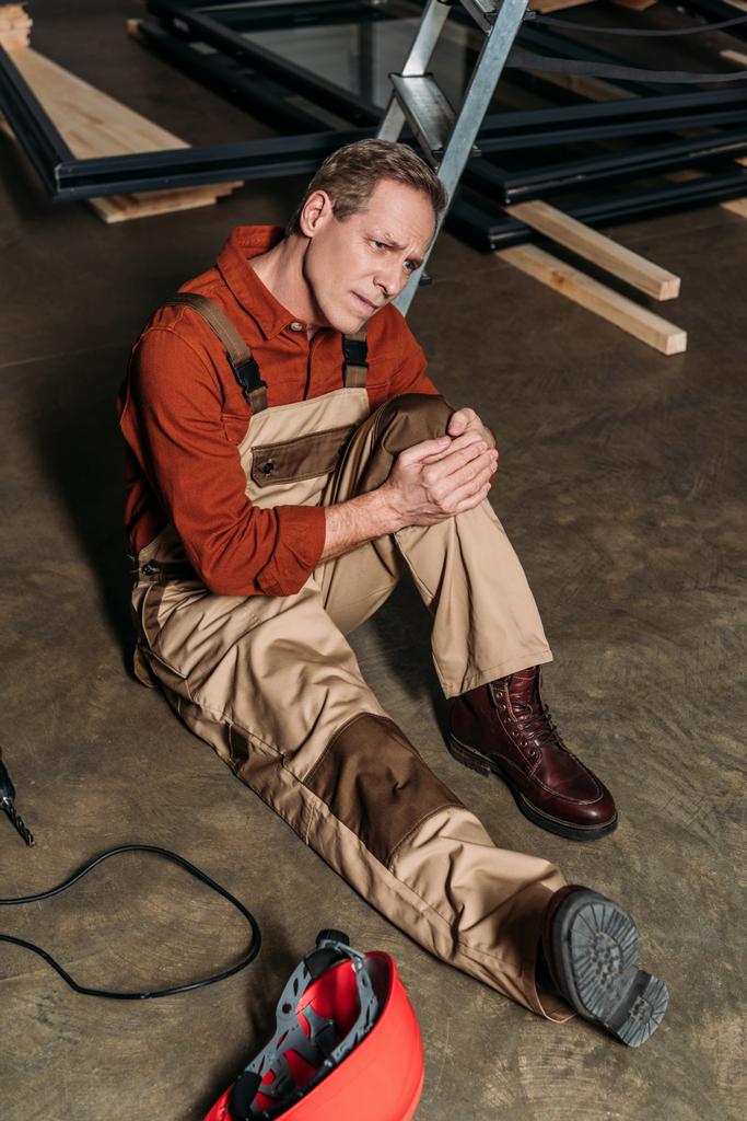 reparador sentado en el suelo y sosteniendo la rodilla lesionada rodeada por el equipo en el garaje
 - Foto, imagen