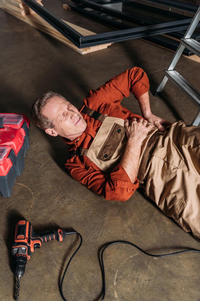 μέσης ηλικίας επισκευαστής ξαπλωμένο στο πάτωμα κοντά Σκάλα και κρατώντας στα πλευρά στο γκαράζ - Φωτογραφία, εικόνα