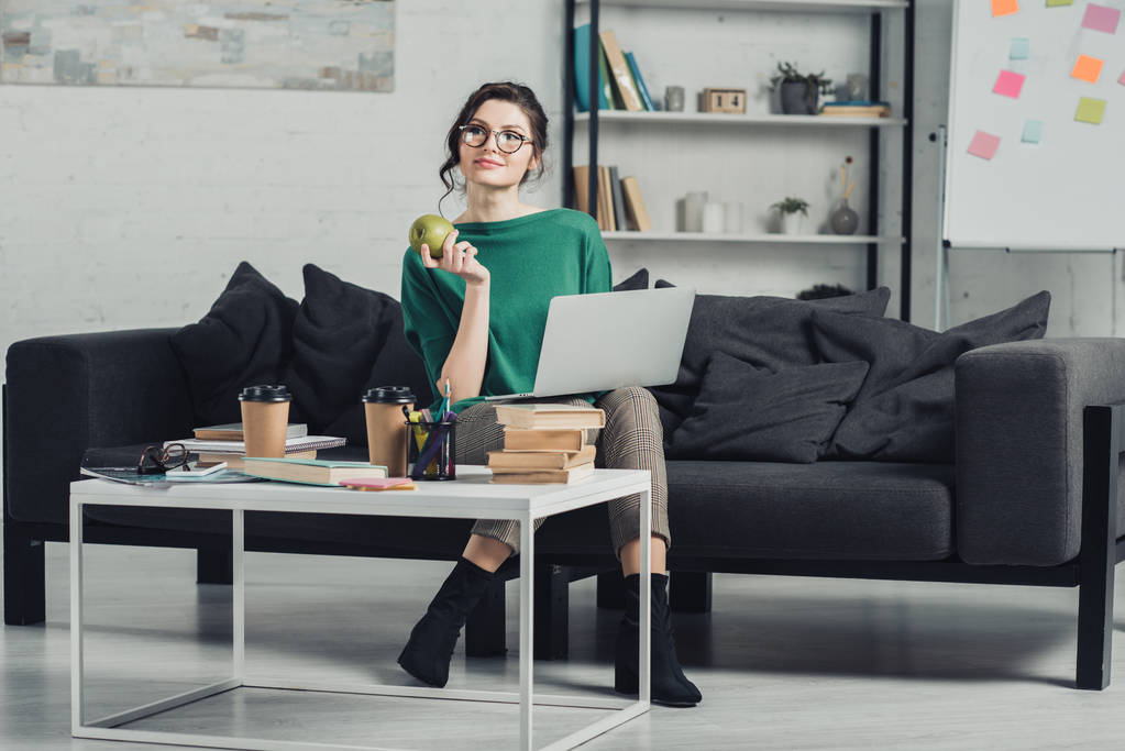 attraente donna in bicchieri che tiene mela mentre seduto con il computer portatile sul divano
 - Foto, immagini