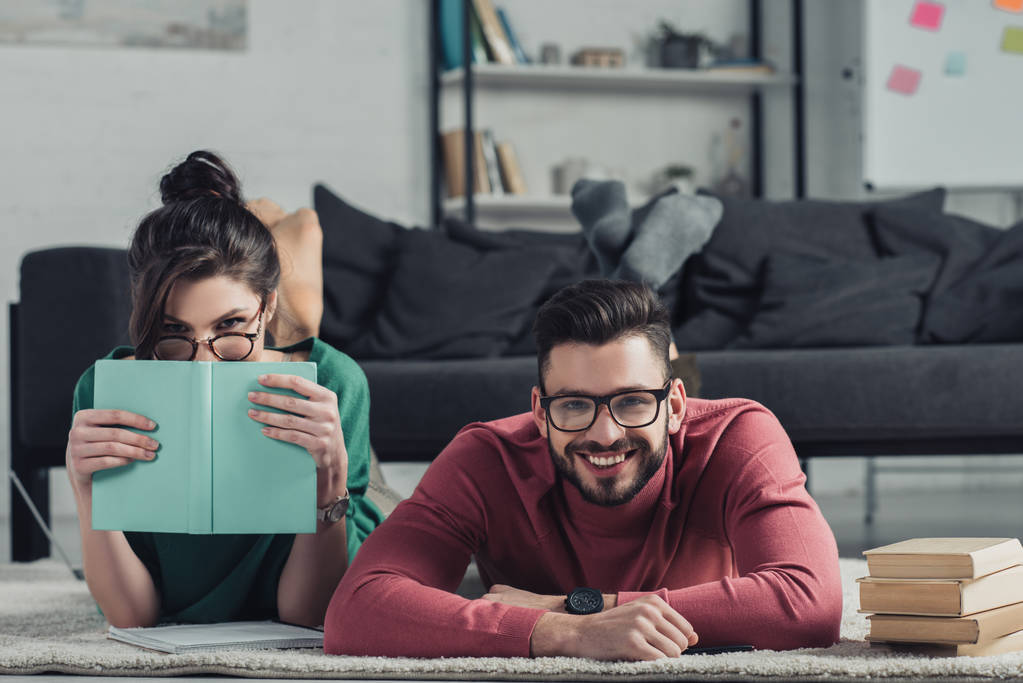 alegre pareja en gafas acostado en la alfombra cerca de libros
  - Foto, Imagen