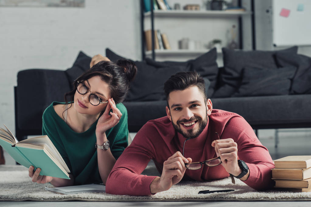 couple joyeux dans des lunettes souriant tout en étant couché sur le tapis avec des livres
  - Photo, image