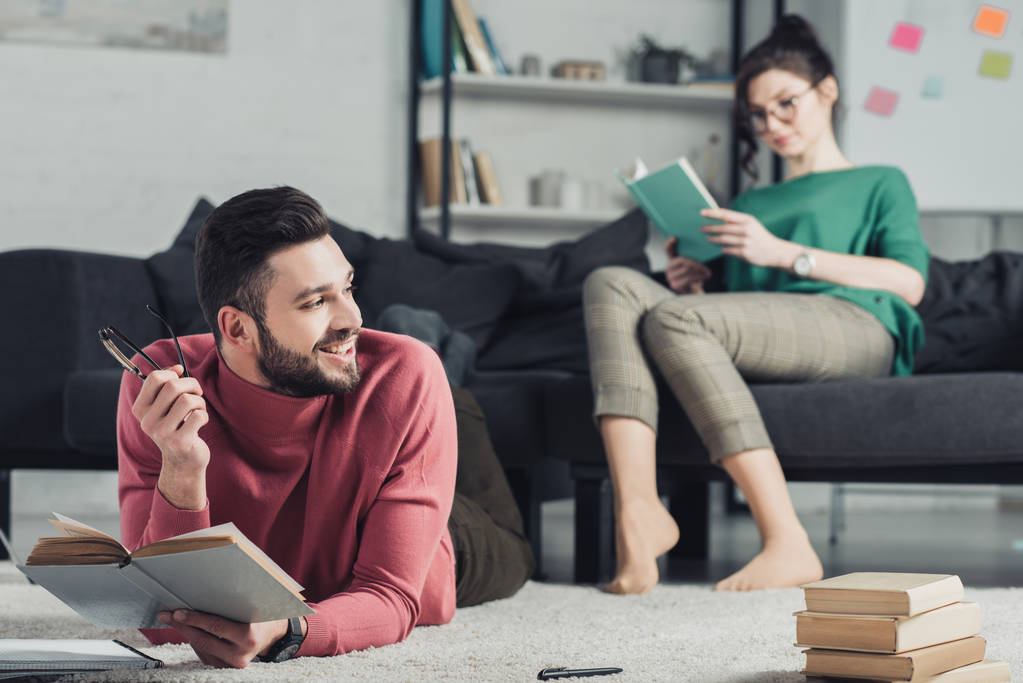 selektiver Fokus des fröhlichen Mannes, der mit Buch studiert, während er auf Teppich liegt, mit Frau im Hintergrund - Foto, Bild