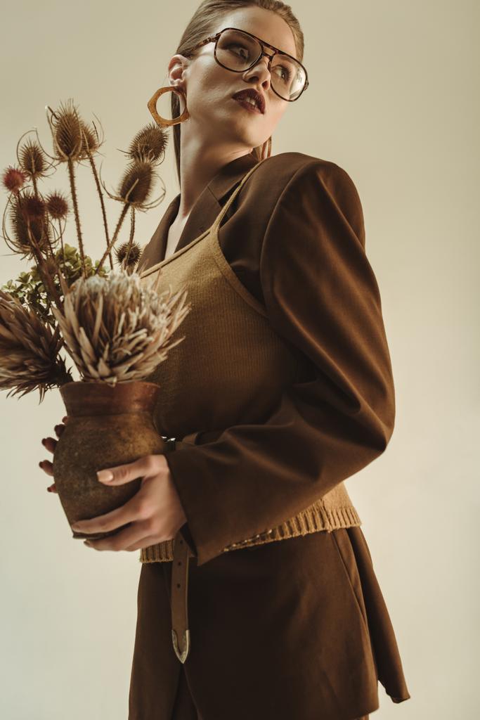 ελκυστική νεαρή γυναίκα κρατώντας πήλινη οινοχόη με μία ανθοδέσμη των ξηρών λουλουδιών που απομονώνονται σε μπεζ - Φωτογραφία, εικόνα