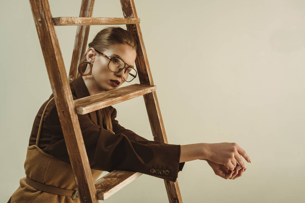 giovane donna alla moda in stile retrò posa vicino scala di legno isolato su beige
 - Foto, immagini