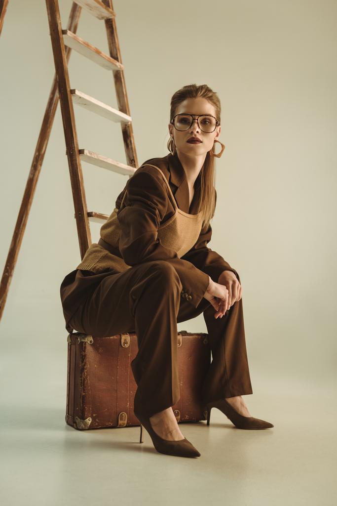 fashionable woman sitting on vintage suitcase near ladder on beige  - Photo, Image