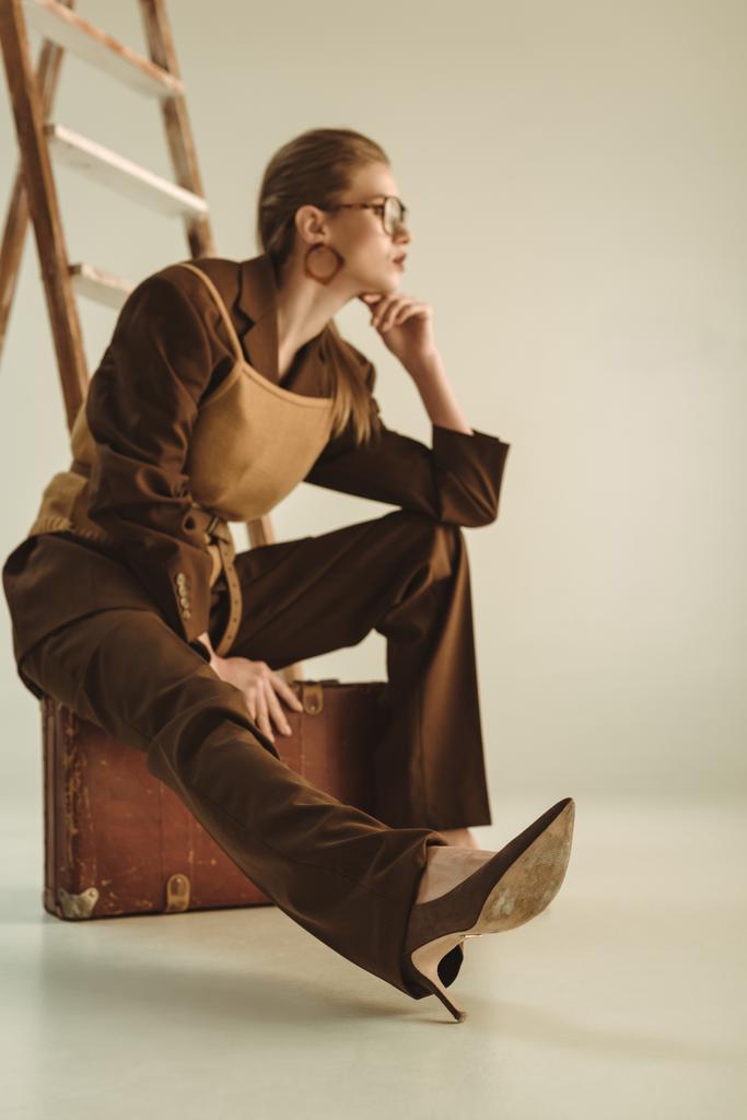 selective focus of stylish girl sitting on vintage suitcase near ladder on beige  - Photo, Image