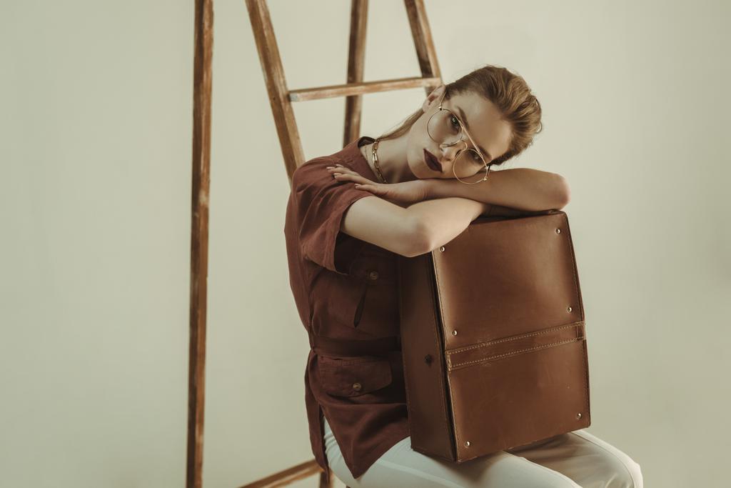 bela jovem segurando saco de couro vintage e sentado perto escada isolada no bege
 - Foto, Imagem