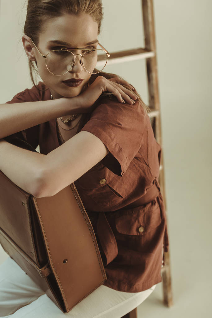 bela jovem segurando saco de couro e sentado perto escada isolada no bege
 - Foto, Imagem