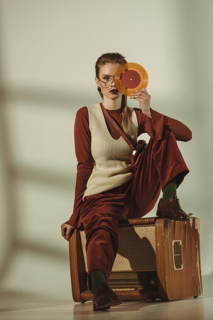 красивая молодая женщина с виниловой пластинкой и сидя на винтажном телевидении на бежевом
 - Фото, изображение