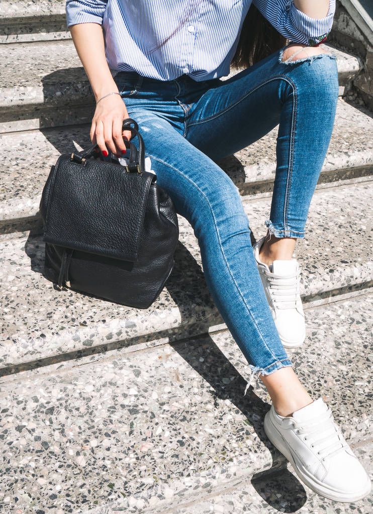 Moda: Una chica en jeans azules se sienta en pasos grises y sostiene una pequeña mochila de cuero de moda
. - Foto, Imagen