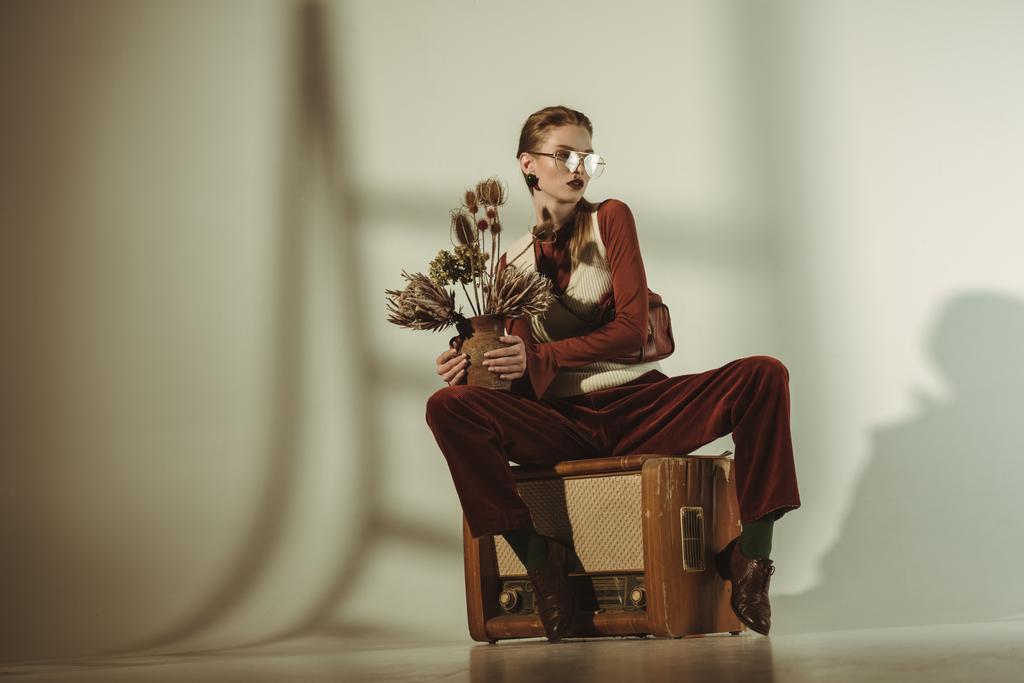 módní holka držení kytice suchých květin a sedí na vintage tv na béžové - Fotografie, Obrázek