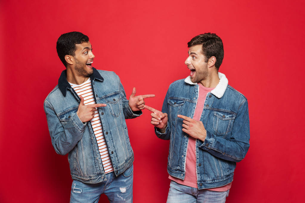 Два взволнованных молодых человека, одетые в джинсовые куртки, стояли изолированные на красном фоне, указывая друг на друга.
 - Фото, изображение