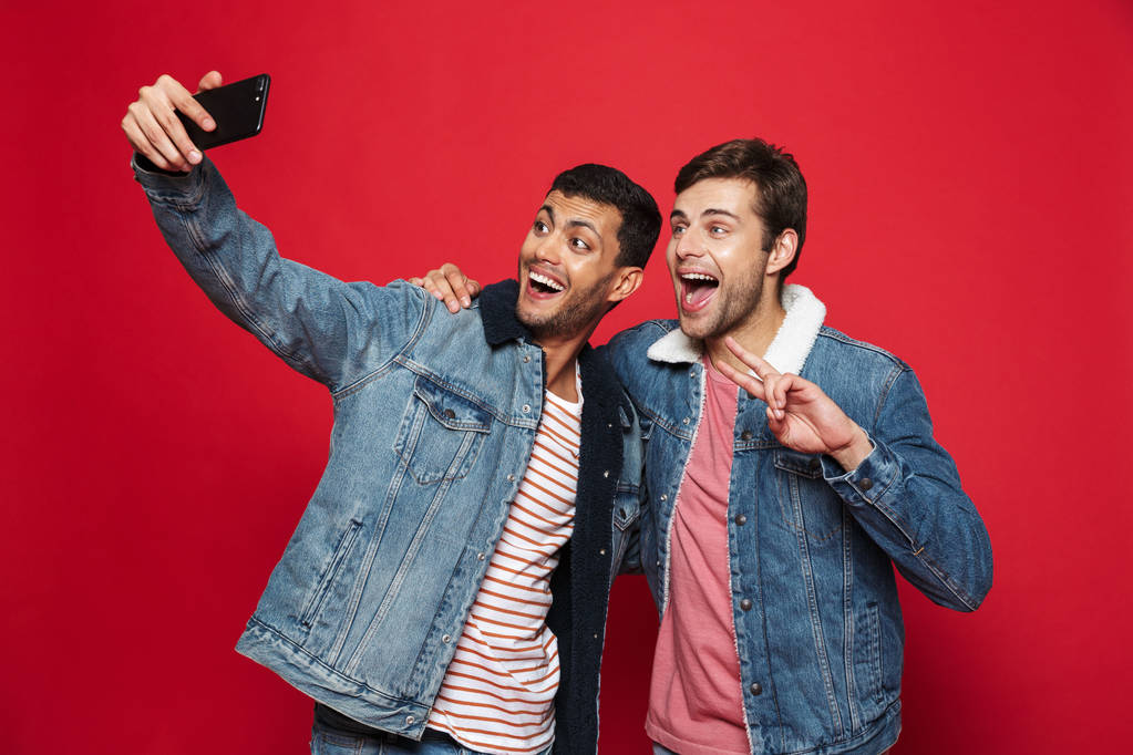 zwei fröhliche junge Männer, die isoliert vor rotem Hintergrund stehen und ein Selfie machen - Foto, Bild