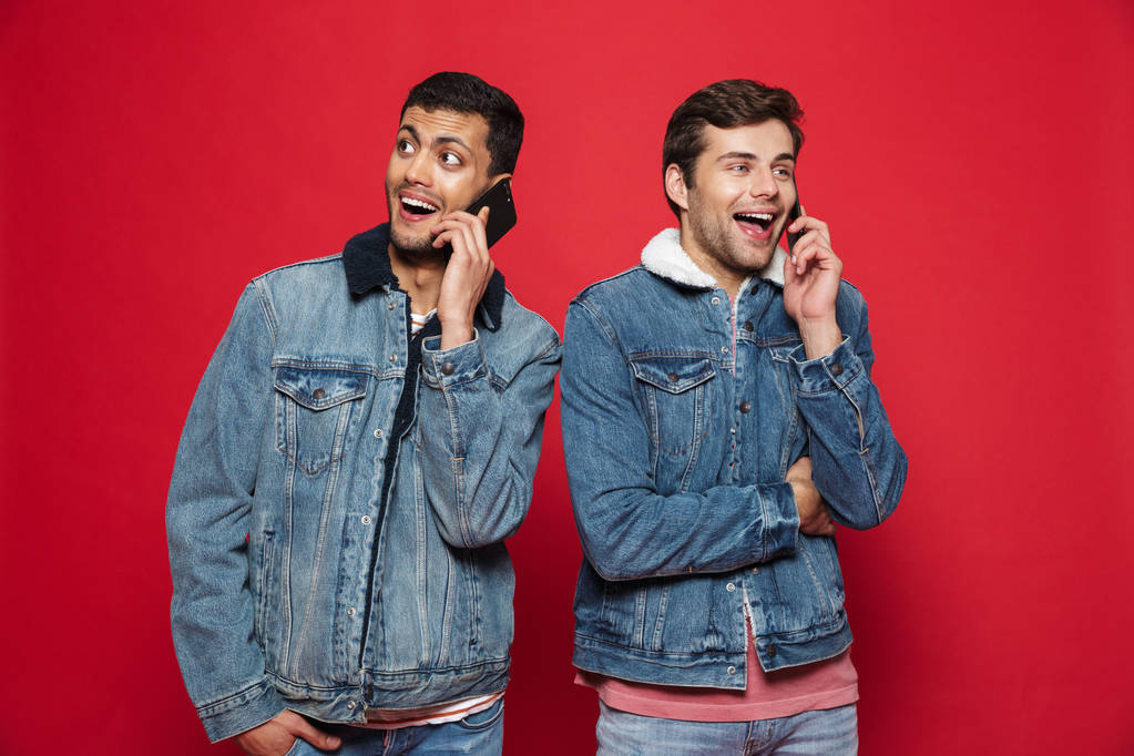 Два веселых молодых человека, стоящих в изоляции на красном фоне, разговаривающих по мобильному телефону
 - Фото, изображение