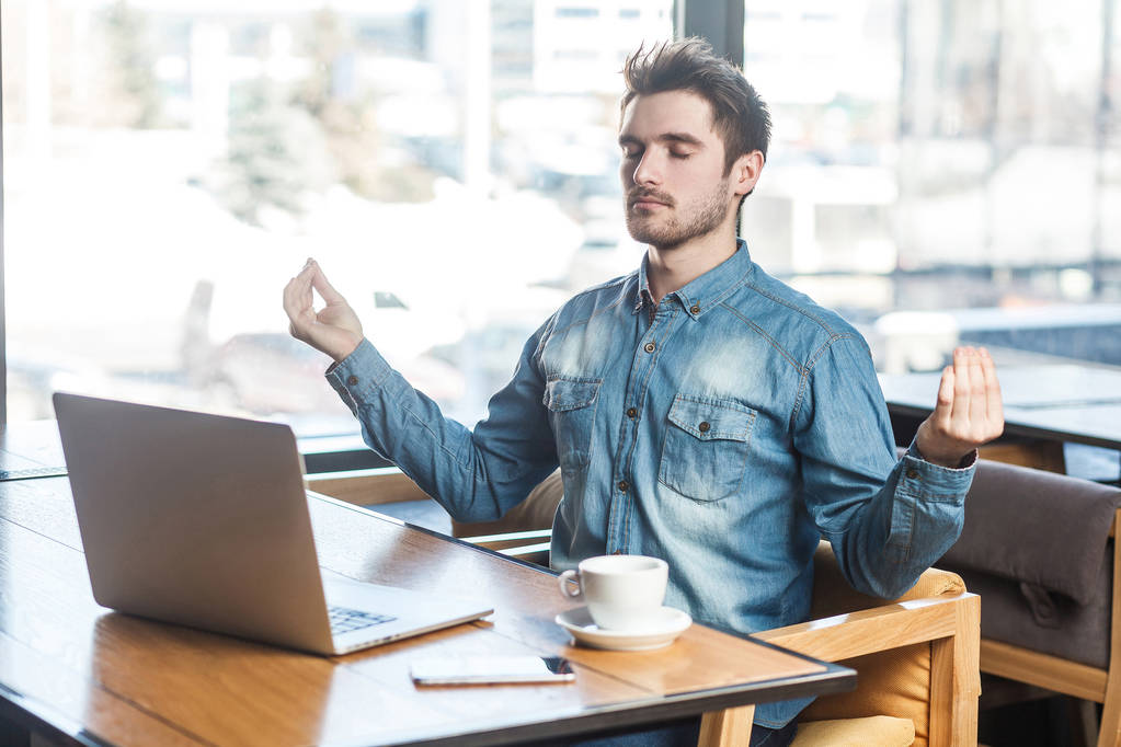 Retrato de belo freelancer de sucesso em camisa jeans azul sentado no café com laptop e tendo descanso ao fazer gesto de ioga
 - Foto, Imagem