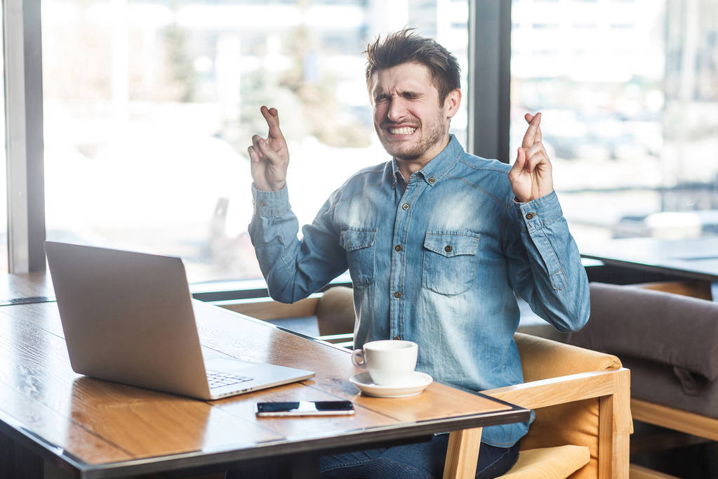 retrato de wishful freelancer em camisa jeans azul sentado com os olhos fechados e dedos cruzados enquanto trabalhava no laptop no café
  - Foto, Imagem