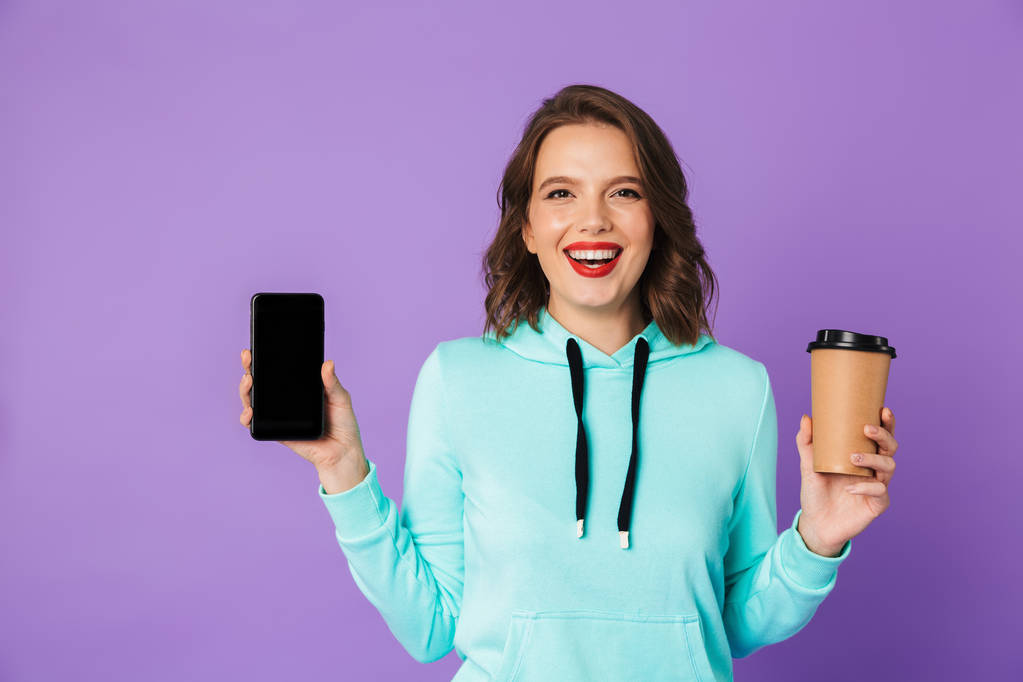 Image d'une belle jeune femme excitée posant isolée sur un mur de fond violet buvant du café montrant l'affichage du téléphone mobile
. - Photo, image